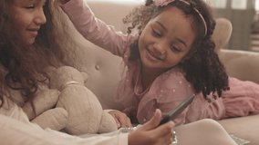 Steadicam shot of curly Biracial little girl wearing pretty outfit, holding toy bear, sitting on couch in living room, her sister lying on sofa, kids smiling and using smartphone