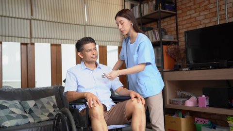 Young Asian female physical therapist standing using a stethoscope put on chest of a middle-aged Asian male patient sitting on a wheelchair. Physical examination while doing therapy treatment at home.