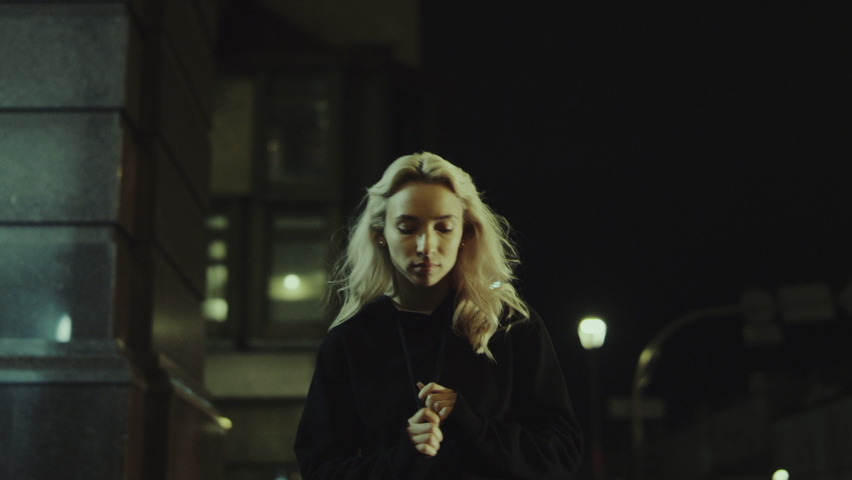 Relaxed woman walking alone while wearing hoodie in night city lights. Blonde girl taking a walk on street at night. Beautiful female person going home late in urban area in slow motion. 