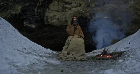 Druid smokes tobacco pipe warming near bonfire against cave