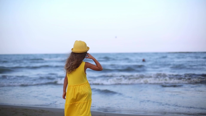 Girl in a yellow dress and hat with long hair fluttering from the breeze looks at the sea and waves, stands with her back to the frame. The end of the vacation, farewell to the sea, summer, nostalgia | Shutterstock HD Video #1085685932