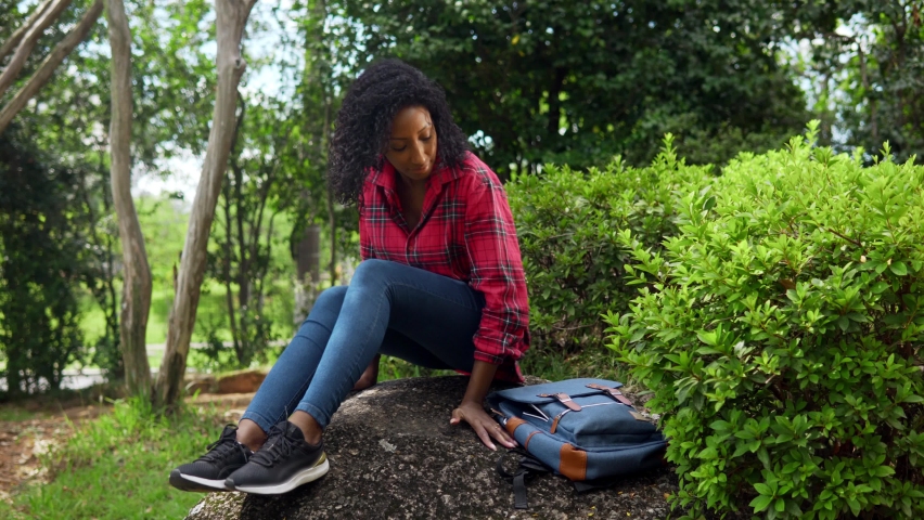 Black quiet woman prepares to read a book of top of a rock in city park.
