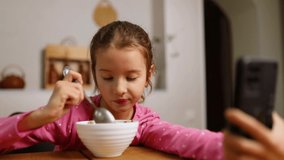 Little girl eating cereal with milk and watching video on smartphone, tasty healthy food, Slow motion of beautiful child having breakfast at home, dependence from gadget