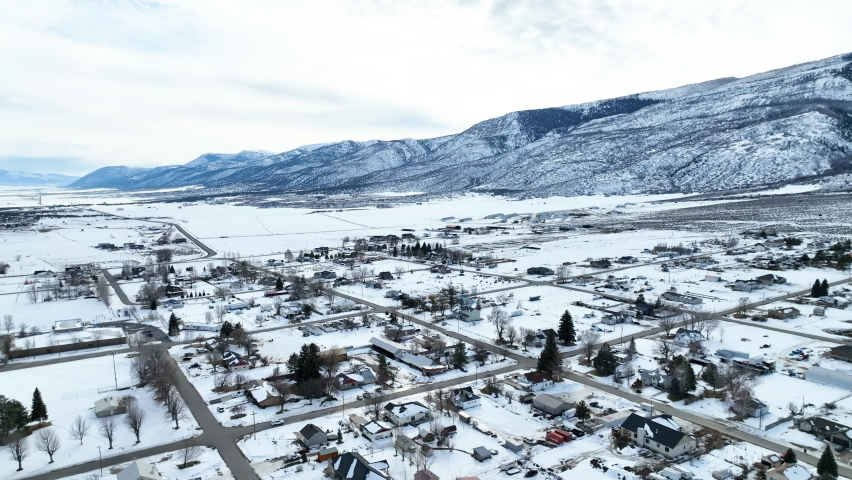 Aerial rural town mountain valley winter snow. Mountain valley cold winter snow agriculture field. Seasonal rural farm city. Homes, business.