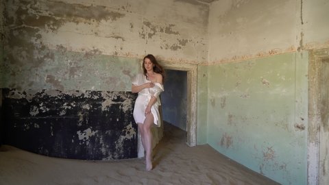 Young woman in robe in abandoned building posing in room. Ghost town Kolmanskop in desert. Room filled with sand in ruined house. Girl in white silk underwear.