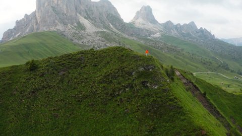 4K Drone video camera flying around person wearing orange jacket on green grass-covered peak in Italian Alps. Environment, hiking, backpacking concept.