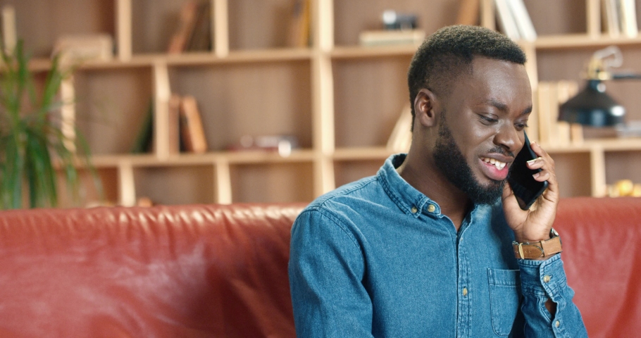 Close up of young african american man in denim shirt sitting on red sofa in modern office and talking on cellphone with client.