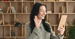 Beautiful Caucasian woman with long dark hair stands on background of bookcase holding tablet and talking with friend on video call.