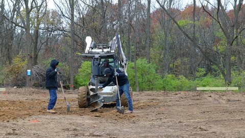 andover , United States - 01 19 2022: Three construction workers using shovels and hydraulic auger mounted on skid steer loader to dig a post hole for the frame of new barn construction;concept of new