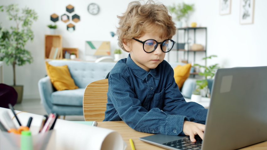 Serious kid in eyeglasses working with laptop computer sitting at desk at home concentrated on online activity. Technology and childhood concept. | Shutterstock HD Video #1085718629