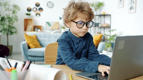 Serious kid in eyeglasses working with laptop computer sitting at desk at home concentrated on online activity. Technology and childhood concept.