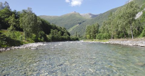Flying Fast through rocky shallow river with crystal white water during sunny day. Mountains in the background. The river surrounded by green trees.
