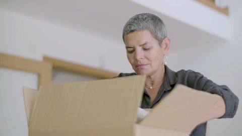 Focused woman unpacking carton boxes after moving to new flat. Cheerful woman with short hair getting things out of cardboard box, enjoying real estate purchase. Relocation, moving concept