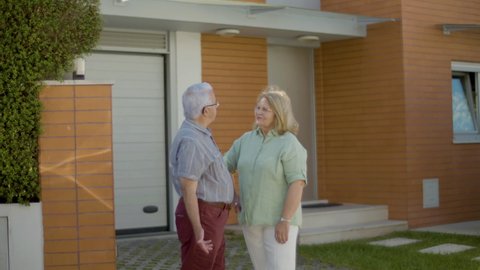 Senior couple standing outdoor and discussing buying new house. Slow motion of beautiful Caucasian man and woman talking after observing apartment on summer day. Real estate purchase concept