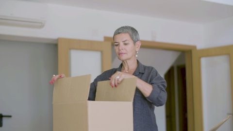 Cheerful woman unpacking carton boxes after moving to new house. Medium shot of beautiful woman with short hair establishing home, rejoicing with real estate purchase. Relocation, moving concept