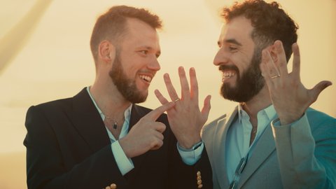 Portrait of a Happy Just Married Handsome Gay Couple in Love Showing Off Their Gold Wedding Rings. Two Attractive Queer Men in Suits Smile and Pose for Camera. LGBTQ Relationship and Family Goals.