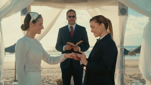 Beautiful Female Queer Couple Exchange Rings and Kiss at Outdoors Wedding Ceremony Near a Sea. Two Lesbian Women in Love Share Their Big Day with Diverse Multiethnic Friends. LGBTQ Relationship Goals.
