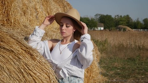 Portrait of sexy lady in hat poses at haystack in summer windy field. 