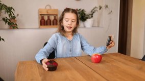 Beautiful young girl filming her blog broadcast about healthy food at the home. Little girl choosing between sweet cake and fresh red apple in , Healthy dieting habit, concept of proper nutrition
