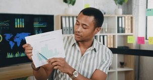 Portrait of successful 30s african man talking and showing sheet of paper with graphs for students looking at laptop at home. Online education process