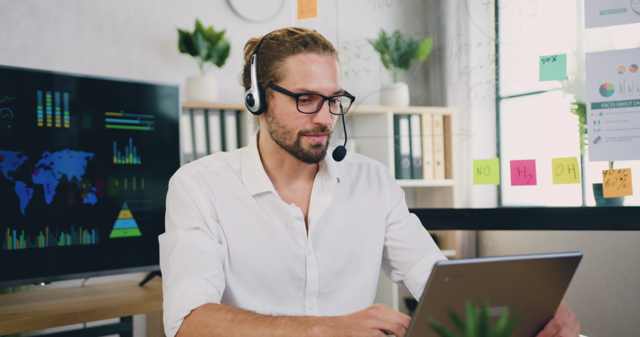 Millennial caucasian male sit at workplace in headset looking at laptop during talking to customer using remote web chat, online video conference application. Royalty-Free Stock Footage #1085741459