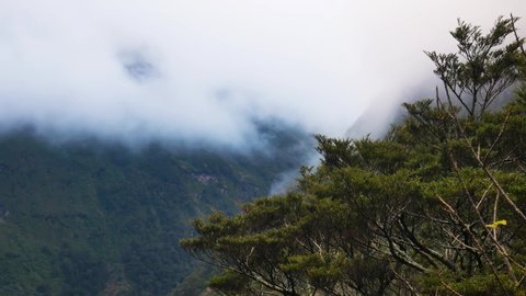 Wide shot of green mountains and rural valley with flying clouds between summits during windy day