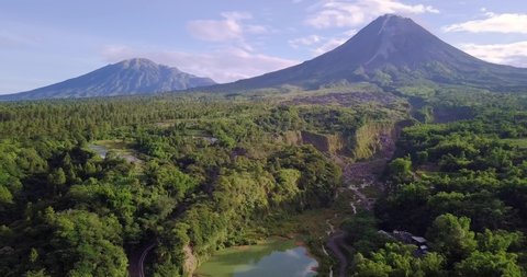 Aerial view of The slopes of Mount Merapi, where there is a river as a route for eruptive material to pass. The vegetation around the river is full of trees. Mount Merapi and Mount Merbabu are visible