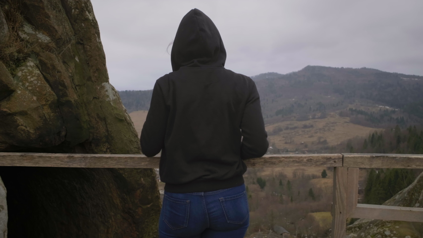 Young adult woman standing in mountains and smiling looking at camera. Female tourist enjoying mountain landscape holding mobile phone in hands.