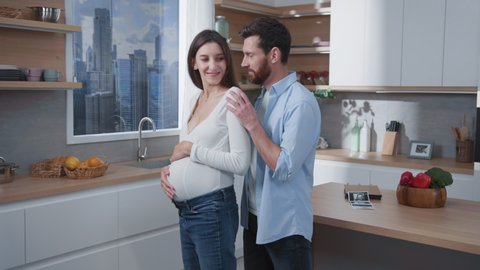 Young loving couple of cheerful bearded caucasian husband with pregnant attractive wife embracing in the kitchen. Lovely family scene. Cozy home. Relationships.