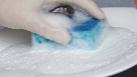 a hand in white surgical glove is holding a blue kitchen sponge with foam for cleaning. liquid for dishes. woman's hand is cleaning white plate. close up video, 4k.