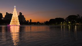 Beautiful christmas tree lit up with warm yellow lights and a star on the tip floating in the waters of the city lake at dusk. Space for text. Video of Igapo lake, Londrina PR Brazil.