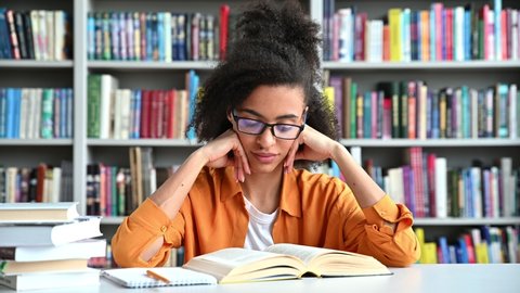 A clever focused african american millennial female student sit in a college library at table, reading book with interest, preparing for lecture or exam, tired and yawns,knowledge and learning concept