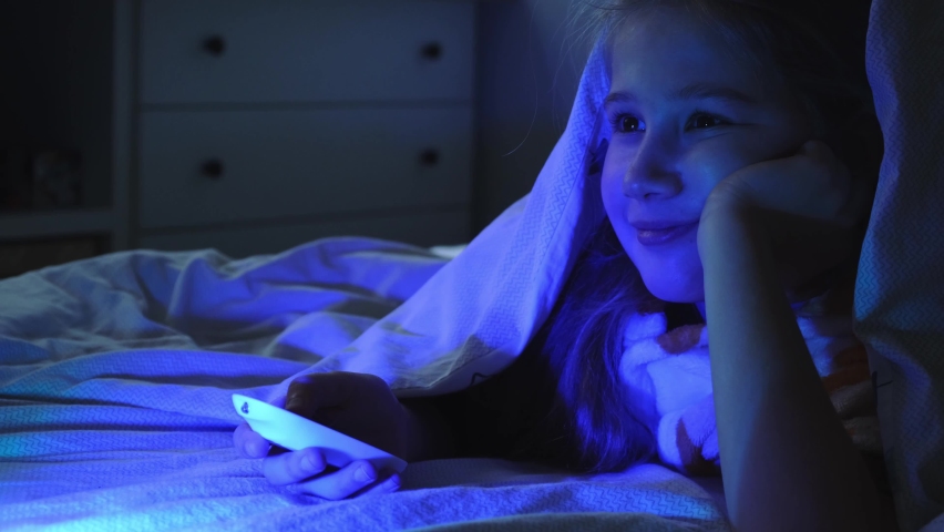 Portrait of cheerful girl while watching TV at night under blanket. Child face illuminated by green and blue neon light from screen. Switching channels with remote. Positive emotions, home life. | Shutterstock HD Video #1085761016