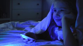Portrait of cheerful girl while watching TV at night under blanket. Child face illuminated by green and blue neon light from screen. Switching channels with remote. Positive emotions, home life.