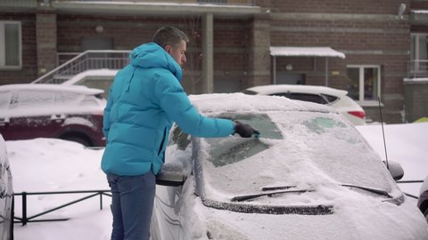 Person driver cleans the car from snow with a brush. A car covered in white snow after a winter snowfall or snow storm. Frozen vehicles in the parking lot.