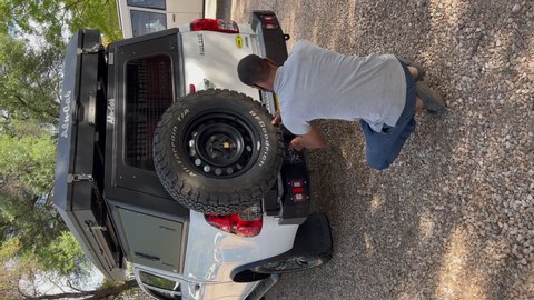 WINDHOEK, NAMIBIA - NOVEMBER, 10, 2021: Vertical video. Black man repairing car, change spare tire at SUV vehicle. Off road white auto with camping equipment for safari offroad travel in Africa.