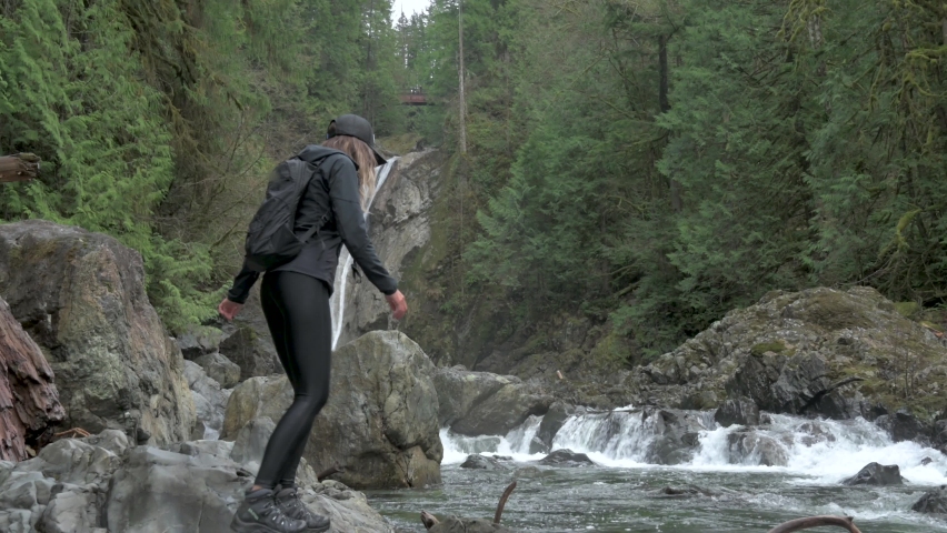 4K slow motion video of young woman hiker jumping from riverbank to a boulder and looking upstream at a plunging waterfall in the Pacific Northwest