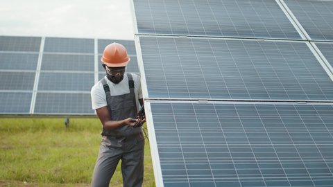 Close up of african american man in grey overalls measuring resistance in solar panels outdoors. Competent technician using multimeter during inspection on station. Man using multimeter for voltage.
