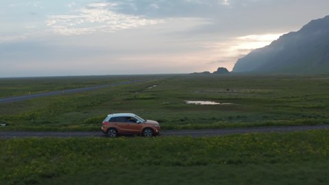 Tracking of SUV car driving on dirt road in countryside at sunrise or sunset. Green flat grassland surrounded by rocky ridge. Iceland
