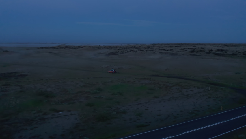 Slide and pan shot of people and car standing in middle of flat plain at twilight. Orbit shot around. Iceland