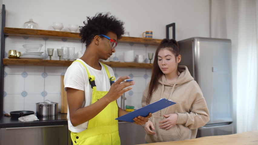 Happy young male plumber holding clipboard for woman signing invoice in kitchen. African-American repairman and Asian female client signing document for services