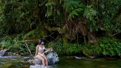 A beautiful woman with long black hair in a white bikini of hispanic ethnicity, is sitting and relaxing on a large rock in a river in nature