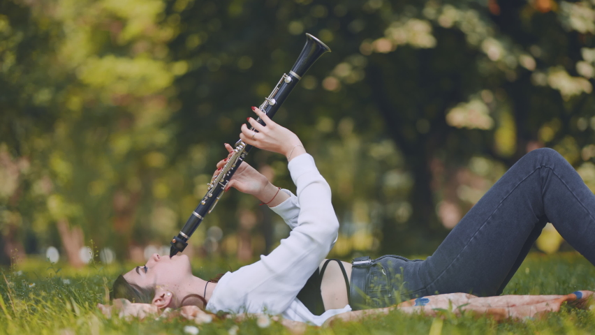 A girl plays the clarinet while lying on the grass in the park.