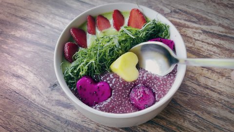 Delicious tropical smoothie bowl with chia seeds, dragon fruit, spinach and strawberry. Dipping out a watermelon heart piece from yummy healthy breakfast with yoghurt and fruits.