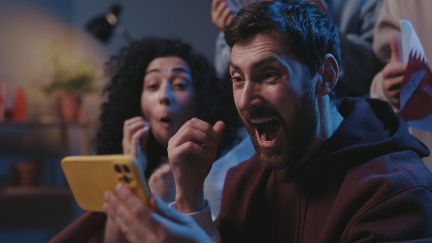 Close up of joyed multiethnic people screaming while watching results of the game on smartphone and sitting on sofa. Focus on bearded guy rejoicing together with friends and screaming