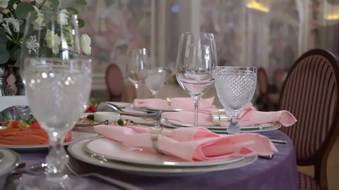 Served table in a restaurant for dinner, lunch or celebration. Purple tablecloth, plates, pink napkin and glasses. Holiday indoors. Empty with no people and food. Decorated for wedding with flowers.