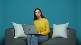 Serious modern young girl in glasses sitting on sofa using laptop, working or learning online, typing on computer