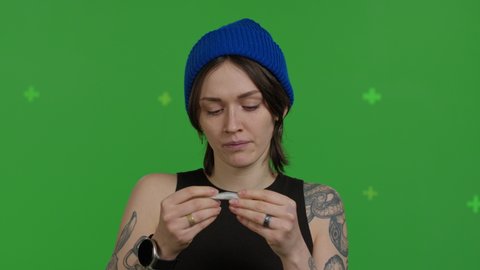 Young sexy female model rolling cannabis joint over green screen background . Pretty girl smoking marijuana addiction on chroma key. 4k raw video footage