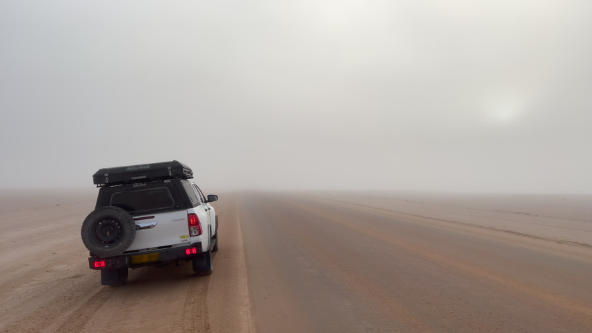 Car at roadside parked in desert. White SUV off road auto vehicle with camping equipment and rooftop tent. Safari travel Africa, Namibia. Gravel dirt road, asphalt highway. Fog in morning. Royalty-Free Stock Footage #1085806220