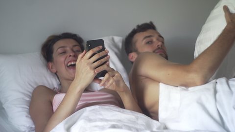 Young Caucasian woman scrolling her phone and laughing loudly in bed, while her husband tries to sleep. Night or morning using phone in bed concept. Phone addict problem.